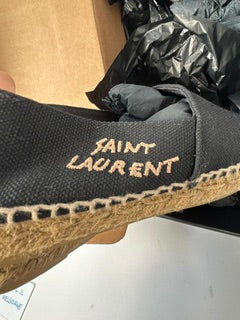 A close-up of a Saint Laurent slip-on shoe in black canvas with tan jute trim and embroidered 'SAINT LAURENT' branding on the side.